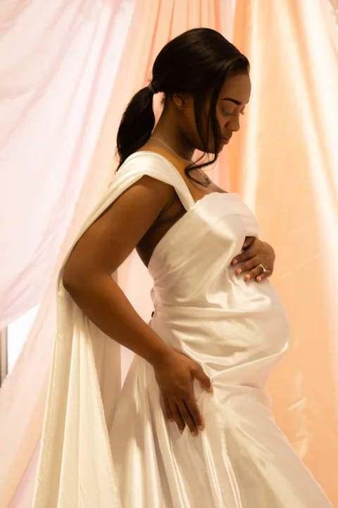 Maternity portrait in a white gown beside a softly lit curtain