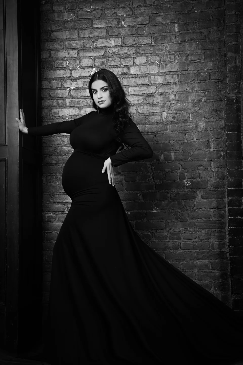 Black-and-white portrait in a long black gown by a brick wall