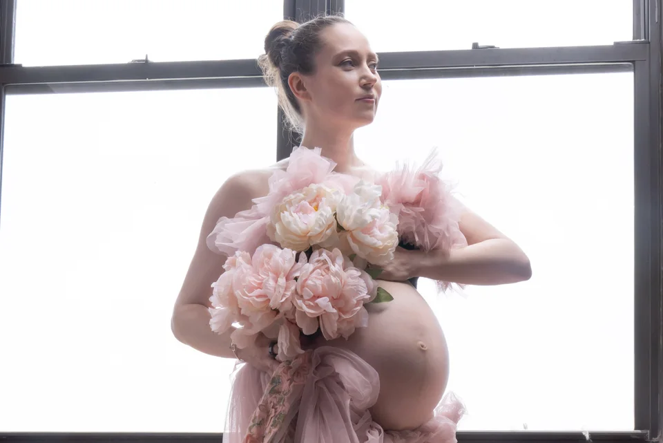 Maternity portrait holding pink flowers beside large windows